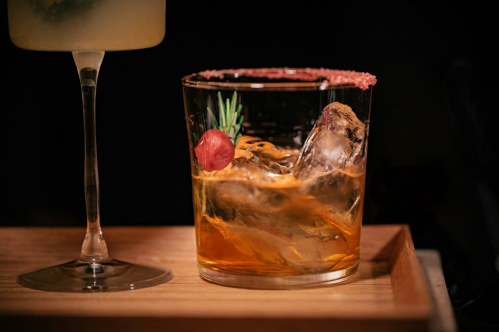 Amber cocktail with ice, cherry and rosemary garnish on a wooden tray.