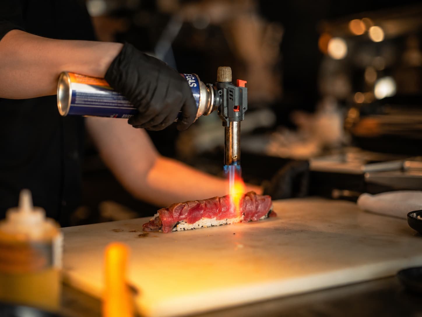 Chef torching aburi sushi with handheld blowtorch in Japanese restaurant kitchen setting.